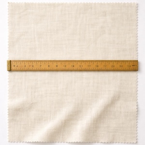 May include: A beige linen fabric sample with a scalloped edge, measuring approximately 24 inches in length, is laid flat. A wooden ruler, marked in inches, is placed across the fabric, providing a visual measurement guide for the material.