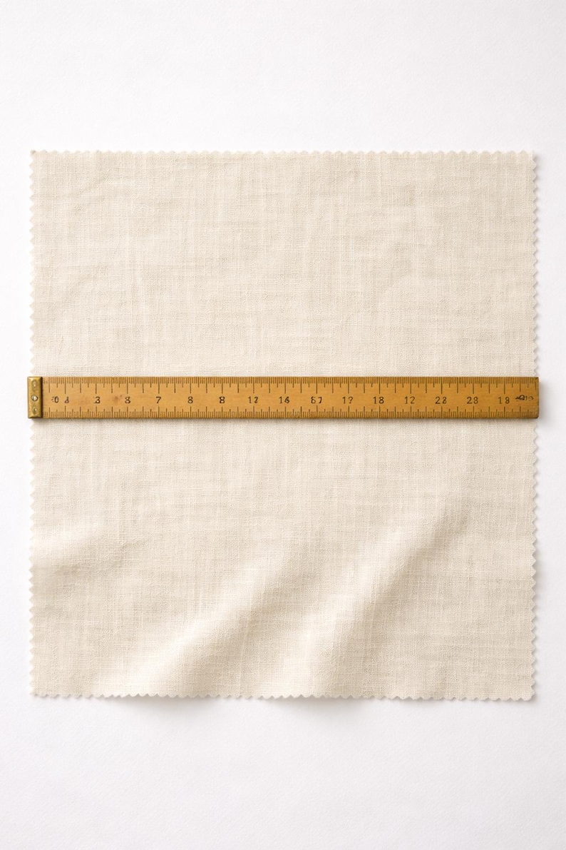 May include: A beige linen fabric sample with a scalloped edge, measuring approximately 24 inches in length, is laid flat. A wooden ruler, marked in inches, is placed across the fabric, providing a visual measurement guide for the material.