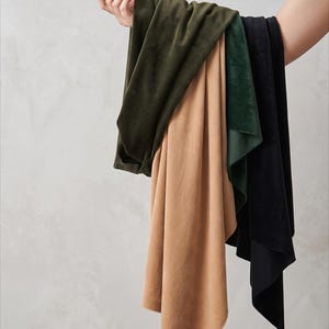May include: A collection of four rectangular fabric swatches in solid colors, including olive green, tan, dark forest green, and black. Each piece of fabric features a soft, short-pile velvet texture that drapes downward in loose, flowing folds.