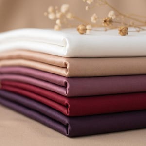 Puede incluir: Una pila de cinco piezas de tela dobladas con un acabado suave y satinado, dispuestas en un degradado desde el blanco en la parte superior hasta el beige, malva, rojo intenso y morado oscuro, rematadas con una ramita de flores secas.