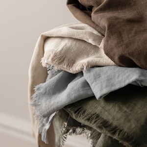 Può includere: Una pila di quattro tessuti piegati con bordi sfrangiati. Il tessuto superiore è marrone scuro, seguito da un tessuto beige chiaro, un tessuto azzurro pallido e un tessuto verde oliva scuro sul fondo. Ogni pezzo presenta una trama visibile.