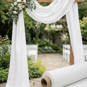 May include: A wooden wedding arch draped with white fabric and adorned with white flowers and greenery. A roll of white fabric sits on the ground. The scene is set outdoors with a pathway and chairs in the background.