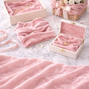 May include: A still-life composition featuring jewelry and floral arrangements. A white box with a gold necklace and a heart pendant, a bracelet, and a basket of white flowers are displayed on a marble surface. Pink velvet fabric adds a soft touch.