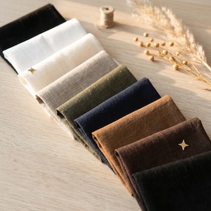 May include: A collection of folded rectangular fabric swatches arranged in a diagonal row, featuring a spectrum of colors including black, white, cream, beige, olive green, navy blue, tan, and dark brown, each accented with a small gold star pin.