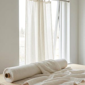 Puede incluir: Un rollo de tela de lino de color natural descansa sobre una mesa de madera clara, con un trozo de tela drapeado sobre el lateral. Cortinas transparentes de color crema cuelgan en el fondo, y unas tijeras descansan sobre la mesa.