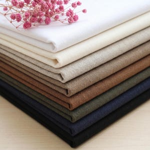 May include: A stack of folded fabric swatches in various neutral colors, including white, beige, brown, and navy. A sprig of pink flowers rests on top of the fabric, adding a touch of color and texture to the display. The fabrics appear to be linen or a similar material.