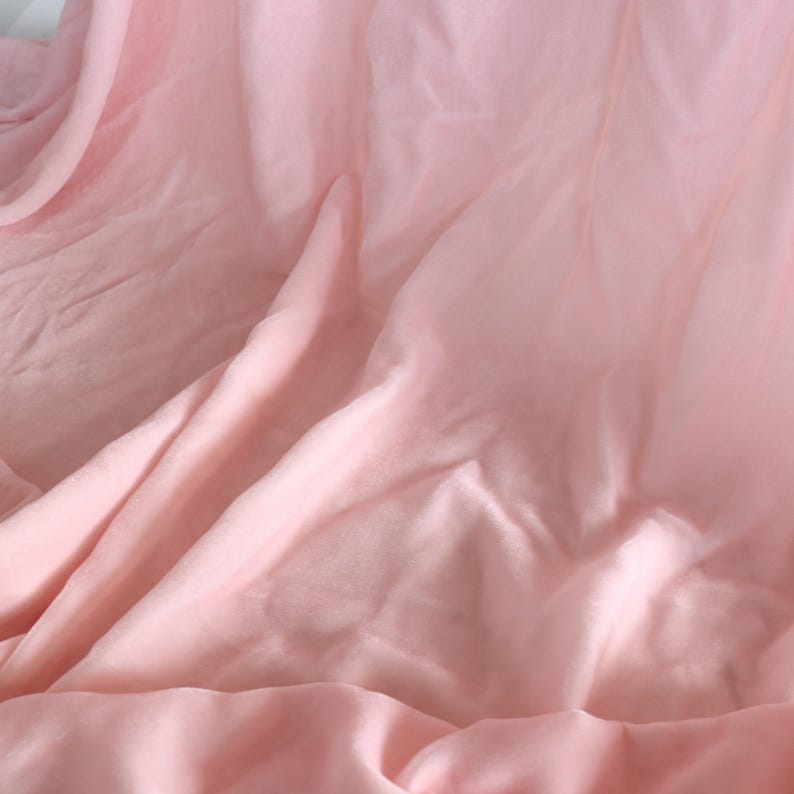 Puede incluir: Una tela de color rosa claro suave con una textura lisa y semitransparente. El material est&aacute; drapeado en pliegues sueltos y fluidos, creando sombras y reflejos sutiles en la superficie. El tejido parece ligero y delicado, con un color rosa pastel uniforme en toda el &aacute;rea visible de la tela.
