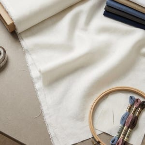 May include: A craft scene featuring a roll of off-white fabric, a wooden embroidery hoop, and spools of colorful embroidery floss. A stack of folded fabric in various shades of blue, beige, and white is also visible.