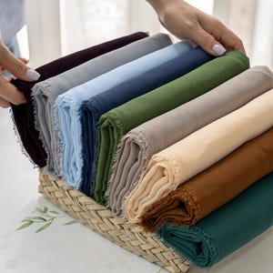 May include: A collection of folded rectangular fabric pieces with frayed edges, arranged in a woven basket. The fabrics are in various solid colors including deep burgundy, gray, light blue, navy, olive green, beige, cream, brown, and dark teal.