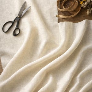 May include: A still-life image of sewing supplies on a wooden surface. The scene includes a pair of black-handled scissors, a beige measuring tape, and a bundle of dried flowers. A piece of cream-colored fabric is draped across the scene.