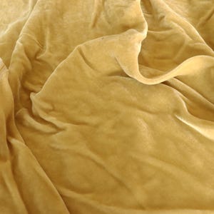 Puede incluir: Una pieza de tela de terciopelo color amarillo mostaza con un pelo denso y suave y un brillo sutil. El material est&aacute; drapeado en pliegues sueltos y fluidos, creando sombras y reflejos profundos en la superficie texturizada del tejido amarillo dorado.