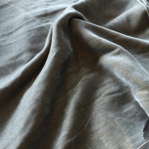 Puede incluir: Una pieza de tela de terciopelo gris oscuro con un pelo suave que refleja la luz. El material est&aacute; drapeado en pliegues sueltos y fluidos, creando sombras y reflejos profundos en la superficie que enfatizan la textura suave y densa del tejido.