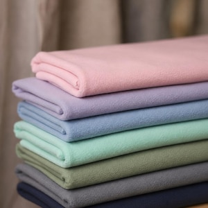 May include: A stack of seven folded pieces of fleece fabric in various solid colors, including light pink, lavender, light blue, mint green, sage green, gray, and navy blue. Each piece of fabric has a soft, matte, and textured surface.