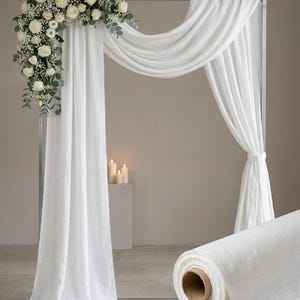 Op de afbeelding: Een witte trouwboog met gedrapeerde stof en een bloemstuk van witte rozen en groen. Een rol witte stof staat op de voorgrond. De boog is gemaakt van een zilveren metalen frame.