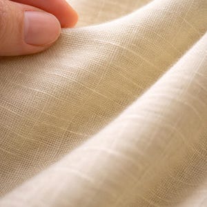 May include: Close-up of a cream-colored fabric with a textured weave. The fabric is folded, creating soft shadows and highlighting the material's subtle variations in color. The texture suggests a natural fiber, possibly linen or cotton.