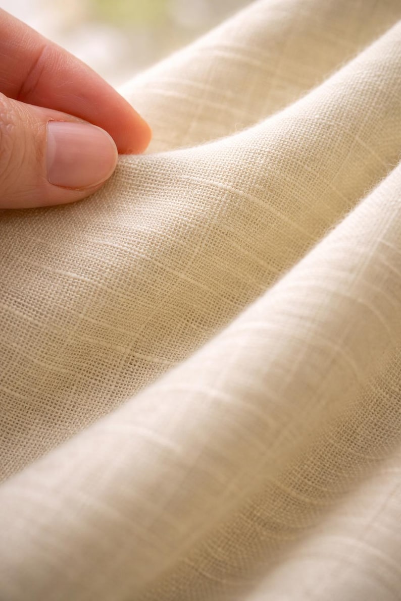 May include: Close-up of a cream-colored fabric with a textured weave. The fabric is folded, creating soft shadows and highlighting the material's subtle variations in color. The texture suggests a natural fiber, possibly linen or cotton.