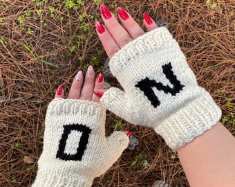Puede incluir: Un par de guantes sin dedos de punto color crema. Cada guante presenta una gran letra negra, uno con la letra "D" y el otro con la letra "N". Los guantes están hechos de un hilo grueso y texturizado.