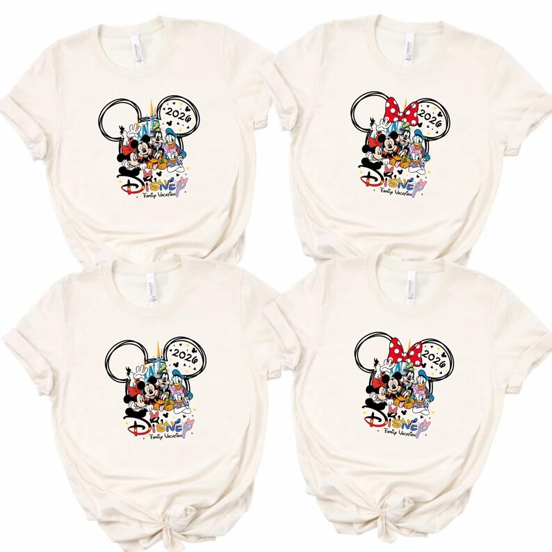 May include: Four cream-coloured t-shirts with a Disney-themed graphic. The design includes Mickey Mouse ears, various Disney characters, the year "2026," and the word "Disney" in a colourful font. One shirt has a red and white polka-dot bow.