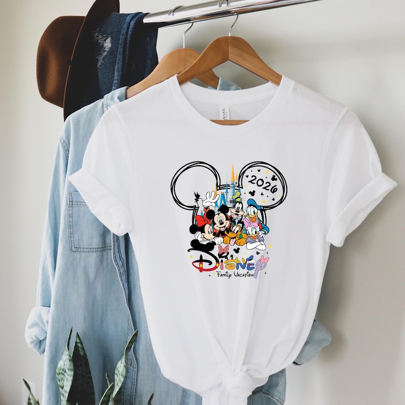 May include: White t-shirt with a Disney-themed graphic. The design includes Mickey Mouse, Minnie Mouse, Donald Duck, and Goofy, with the text "Disney Family Vacation" and the year "2026" inside a Mickey Mouse head outline.