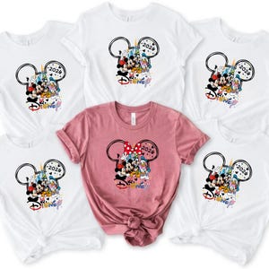 May include: White and pink t-shirts with a Disney design. The design features Mickey Mouse, Minnie Mouse, Donald Duck, and other characters, with the year 2026. The word "Disney" is also present in a colourful font.