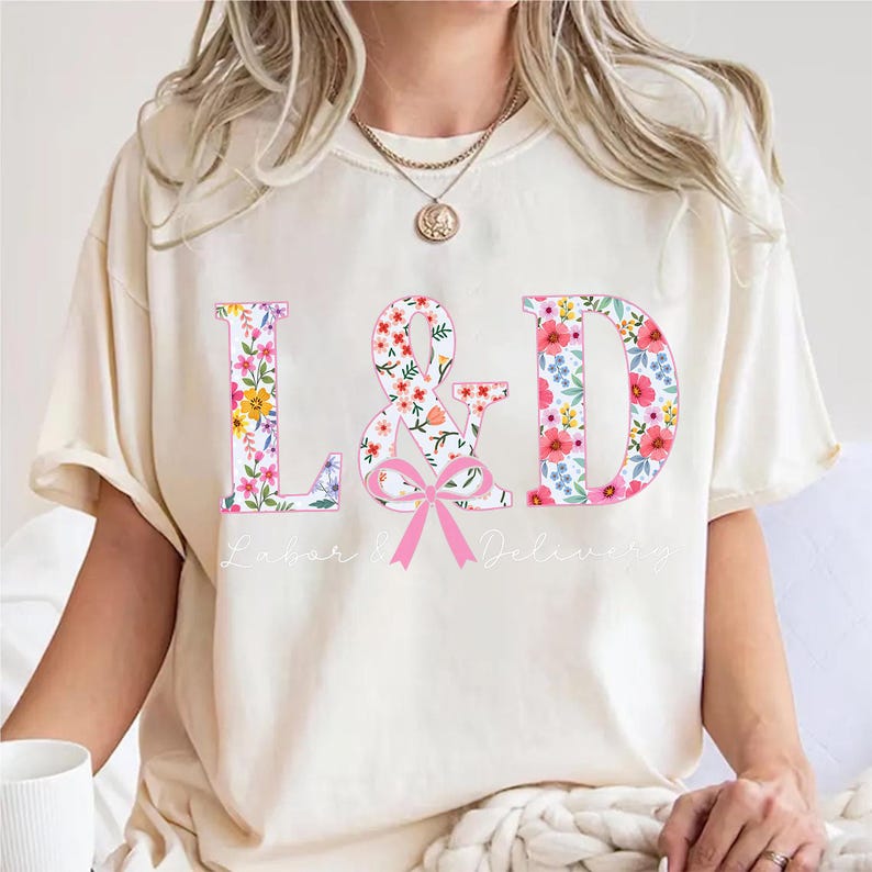 Puede incluir: Camiseta color crema con las letras "L&D" en un dise&ntilde;o floral. El s&iacute;mbolo "&" y las letras est&aacute;n llenos de flores coloridas. Debajo del s&iacute;mbolo "&" hay un lazo rosa, y debajo est&aacute;n impresas las palabras "Labor & Delivery".