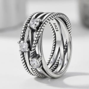 May include: A silver-coloured ring featuring a multi-band design with twisted rope-like textures and three round, clear, prong-set stones. The inner band is clearly engraved with the hallmark S925, indicating the metal composition of the jewellery piece.