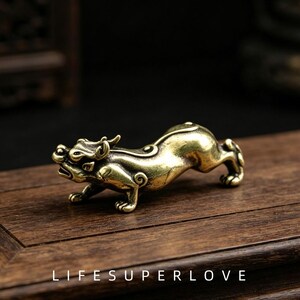 Puede incluir: Una pequeña figura de latón en tono dorado que representa a una criatura mítica en posición agachada y saltarina. La criatura presenta cuernos estilizados, una espalda curva y patas musculosas detalladas. El texto LIFESUPERLOVE está impreso debajo.