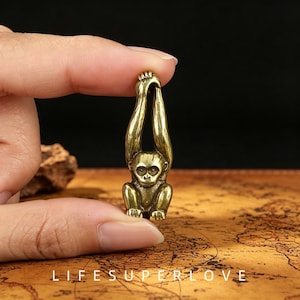 Peut inclure: Une petite figurine de singe en laiton doré métallique représentant un primate assis avec les bras tendus vers le haut, formant une boucle au sommet. Le singe présente des traits faciaux détaillés, incluant des yeux, un nez et une bouche.