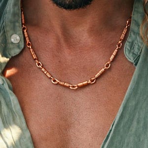 May include: A copper-colored necklace with a unique design. The necklace features a series of cylindrical and coiled elements connected by small links. The necklace is worn over a light green button-down shirt.
