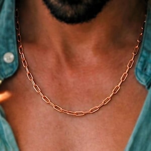 May include: A rose gold chain necklace featuring elongated, rectangular links. The necklace rests against the neck, showcasing a metallic finish and a uniform, repeating link pattern that creates a continuous, structured design around the collar.