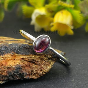 Könnte beinhalten: Ein silberner Ring mit einem tiefroten, ovalen Edelstein. Der Ring liegt auf einer dunklen, strukturierten Oberfläche mit einem verschwommenen Hintergrund aus gelbem und grünem Laub. Das Design des Rings ist schlicht und elegant.