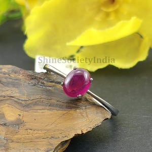 Könnte beinhalten: Ein silberner Ring mit einem großen, ovalen, tiefroten Edelstein. Der Ring liegt auf einer dunklen, strukturierten Oberfläche, mit leuchtend gelben Blumen im Hintergrund. Der Ring ist ein Schmuckstück.