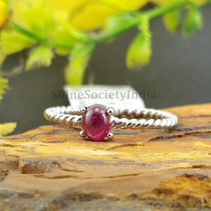 Könnte beinhalten: Ein silberner Ring mit einem gedrehten Band und einem leuchtenden, oval geformten Rubin-Edelstein. Der Ring liegt auf einer natürlichen Holzoberfläche mit einem unscharfen Hintergrund aus gelben Blumen und Grün. Der Ring ist ein Schmuckstück.