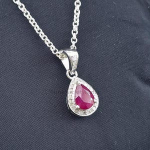 Può includere: Una collana con pendente in argento con una pietra preziosa rosso scuro a forma di goccia, circondata da piccole pietre trasparenti. Il pendente è appeso a una delicata catena d'argento. La collana è mostrata su uno sfondo scuro.