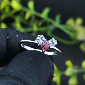 Könnte beinhalten: Ein silberner Ring mit drei quadratischen Edelsteinen. Der zentrale Stein ist tiefrot, flankiert von zwei helleren, quadratisch geschliffenen Steinen. Der Ring ist vor einem dunklen Hintergrund mit grünem Laubwerk platziert.