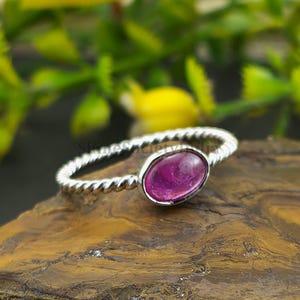 Könnte beinhalten: Ein Silberring mit gedrehtem Band und einem einzelnen ovalen, dunkelrosa Edelstein in einer glatten Metallfassung. Der Ring liegt auf einer strukturierten, braunen, geschichteten Steinoberfläche und präsentiert den Metallglanz und den Stein.