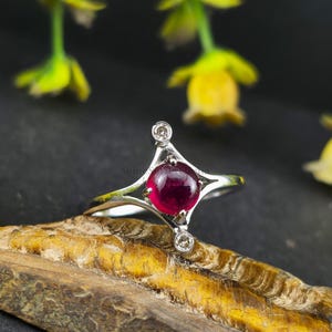 Könnte beinhalten: Ein silberner Ring mit Sternmuster, mit einem runden, tiefroten Edelstein in der Mitte. Zwei kleinere, klare Edelsteine sind an den Spitzen des Rings gesetzt. Der Ring wird auf einer Holzoberfläche mit gelben Blumen im Hintergrund präsentiert.