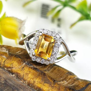 Könnte beinhalten: Ein silberner Ring mit einem rechteckigen, gelben Edelstein in der Mitte, umgeben von kleineren, klaren Steinen. Das Band des Rings hat ein verdrehtes Design. Der Ring liegt auf einer braunen, strukturierten Oberfläche.