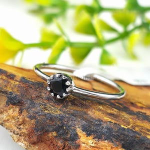 Könnte beinhalten: Ein silberner Ring mit einem kleinen, runden, schwarzen Edelstein. Der Ring liegt auf einem Holzstück mit natürlicher, strukturierter Oberfläche. Grünes Laub ist im Hintergrund zu sehen.