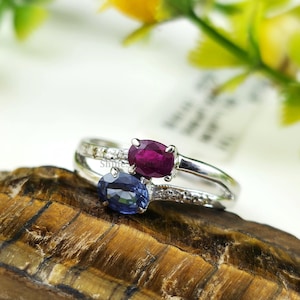 Könnte beinhalten: Ein Silberring mit einem tiefroten Rubin und einem blauen Saphir. Der Ring hat ein geteiltes Band mit kleinen Akzentsteinen. Der Ring wird auf einer strukturierten, braunen Oberfläche mit einem verschwommenen Hintergrund aus Grünzeug präsentiert.