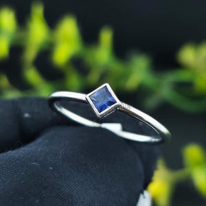 Könnte beinhalten: Ein silberner Ring mit einem quadratischen, tiefblauen Edelstein. Der Ring hat ein schmales Band und ein minimalistisches Design. Der Edelstein ist in einer quadratischen Lünette gefasst, was einen modernen und eleganten Look erzeugt. Der Ring wird vor einem dunklen Hintergrund mit grünem Laub gezeigt.