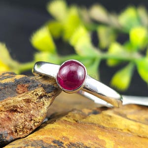 Könnte beinhalten: Ein Silberring mit einem großen, ovalen, tiefroten Edelstein. Das Band des Rings ist glatt und poliert, und der Edelstein ist in einer einfachen Lünettenfassung gefasst. Der Ring wird auf einer strukturierten, braunen und gelben Oberfläche präsentiert.