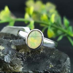 Könnte beinhalten: Ein silberner Ring mit einem ovalen Opal-Edelstein. Der Opal hat eine hellgelbe und weiße Farbe. Der Ring liegt auf einer dunklen, strukturierten Oberfläche mit grünem Laub im Hintergrund.