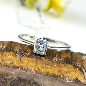 Könnte beinhalten: Ein silberner Ring mit einem rechteckigen, hellblauen Edelstein. Der Edelstein ist in einem quadratischen Rahmen gefasst. Der Ring wird auf einer strukturierten, braunen Oberfläche präsentiert. Es ist ein Schmuckstück.