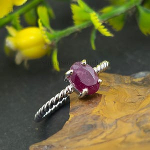 Könnte beinhalten: Ein silberner Ring mit einem gedrehten Band und einem großen, oval geformten Rubin-Edelstein. Der Rubin hat eine tiefrote Farbe und ist in einer Vier-Zacken-Fassung gefasst. Der Ring wird auf einer braunen und gelben Oberfläche mit grünem Laub im Hintergrund präsentiert.