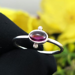 Könnte beinhalten: Ein silberner Ring mit einem tiefvioletten, ovalen Edelstein, flankiert von zwei kleinen, runden, klaren Steinen. Der Ring ist vor einem verschwommenen Hintergrund aus gelben Blumen und einer schwarzen Oberfläche platziert.