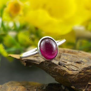 Può includere: Un anello d'argento con una grande gemma ovale di colore rosso intenso. La gemma è incastonata in una semplice fascia d'argento lucida. L'anello è esposto su uno sfondo di fiori gialli e una superficie di legno.