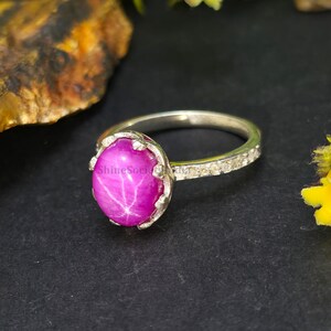 Könnte beinhalten: Ein silberner Ring mit einem großen, oval geschliffenen Sternrubin. Der Rubin hat eine leuchtend rosa Farbe mit einem sternförmigen Effekt. Der Ring ist mit kleinen, klaren Edelsteinen besetzt. Der Ring befindet sich vor einem dunklen Hintergrund.