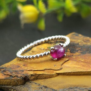 May include: A silver ring featuring a twisted band design, set with a large, round, magenta gemstone. The ring is displayed on a textured stone surface, with yellow flowers in the background.