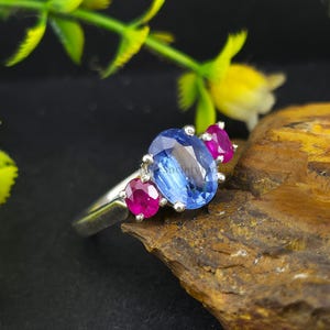 May include: A silver ring featuring a large, oval-cut blue gemstone flanked by two smaller, round-cut pink gemstones. The ring is set on a wooden surface with a dark background, and is accented by green and yellow foliage.